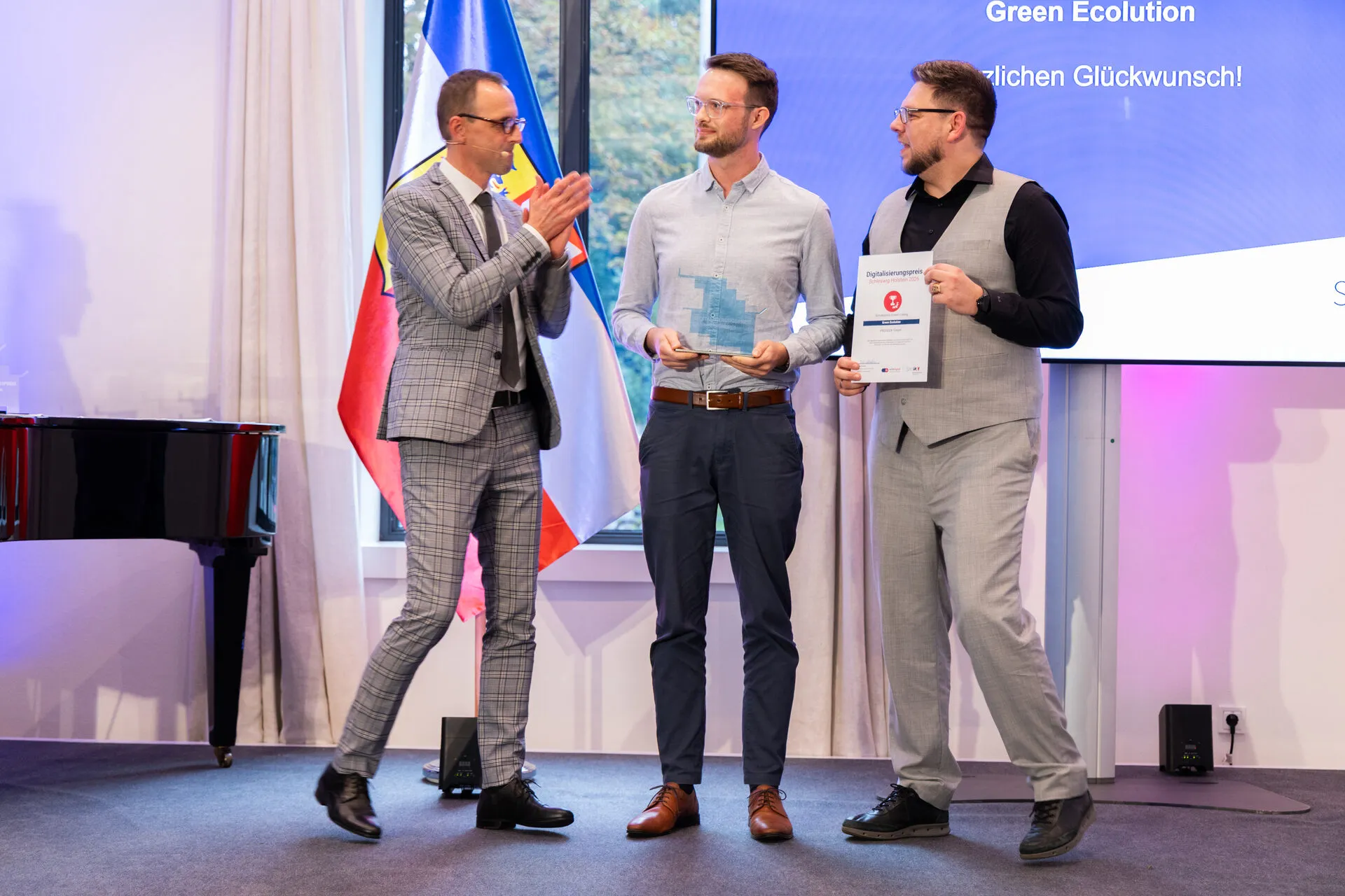 Digitalisierungsminister Dirk Schrödter applaudiert auf der Bühne zwei Vertretern von PROGEEK mit Trophäe und Urkunde, dahinter die Leinwand mit der Aufschrift Green Ecolution