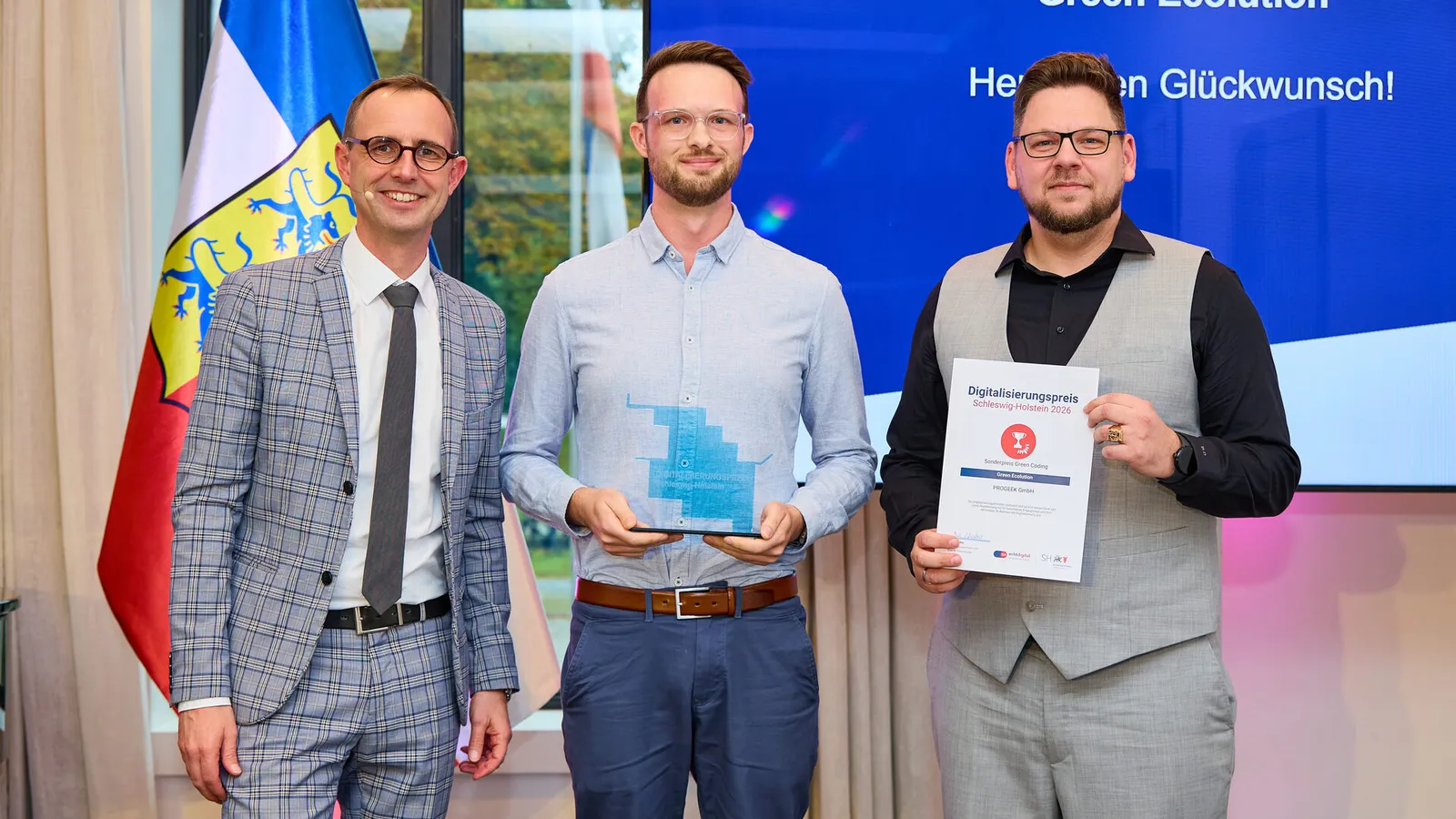 Digitalisierungsminister Dirk Schrödter steht mit zwei Vertretern von PROGEEK vor einer Leinwand mit der Aufschrift „Green Ecolution, Herzlichen Glückwunsch!“, einer hält die gläserne Trophäe, der andere die Urkunde