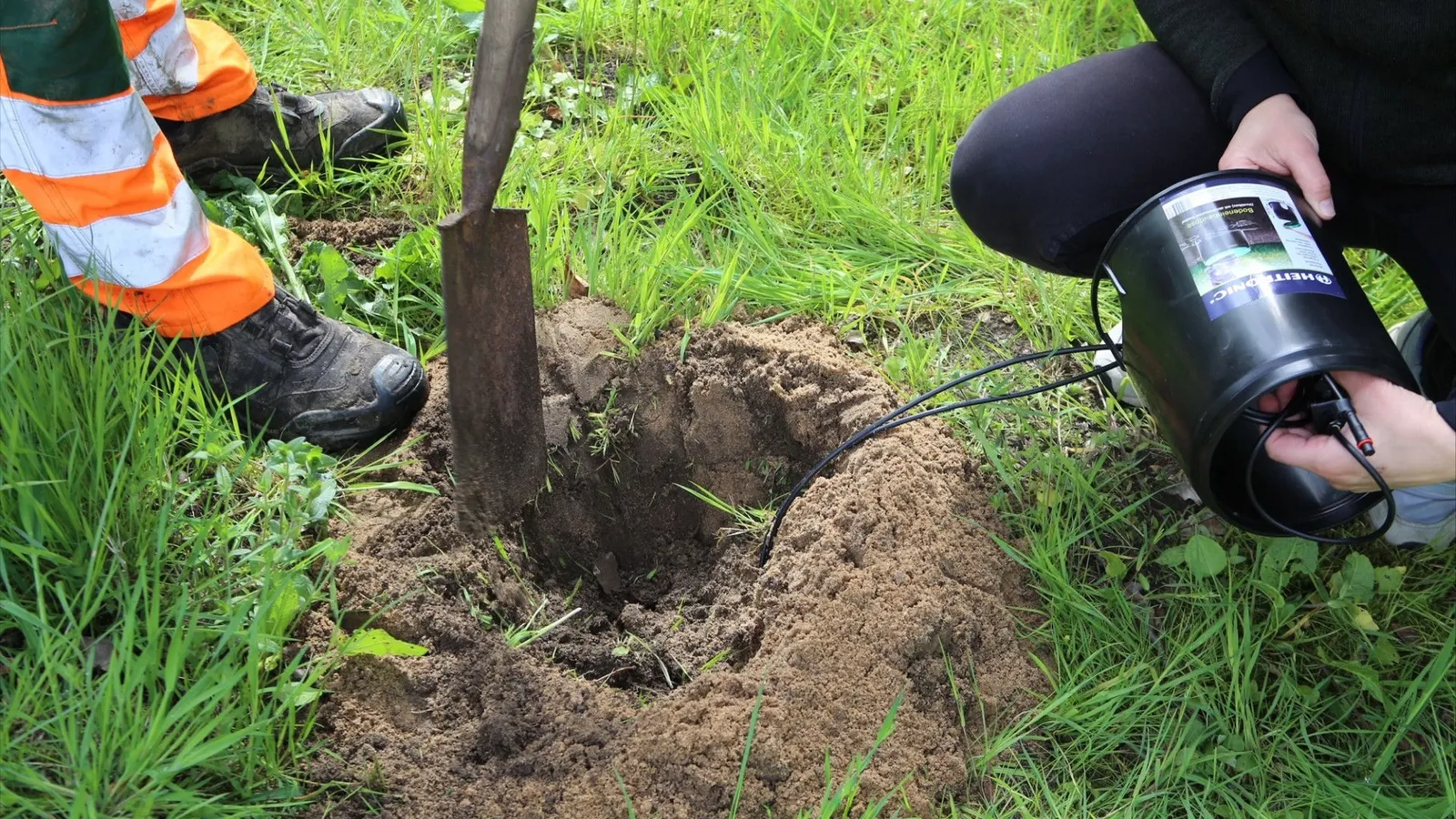 Two people place a sensor unit into a freshly dug hole, next to it a spade stuck in the grass