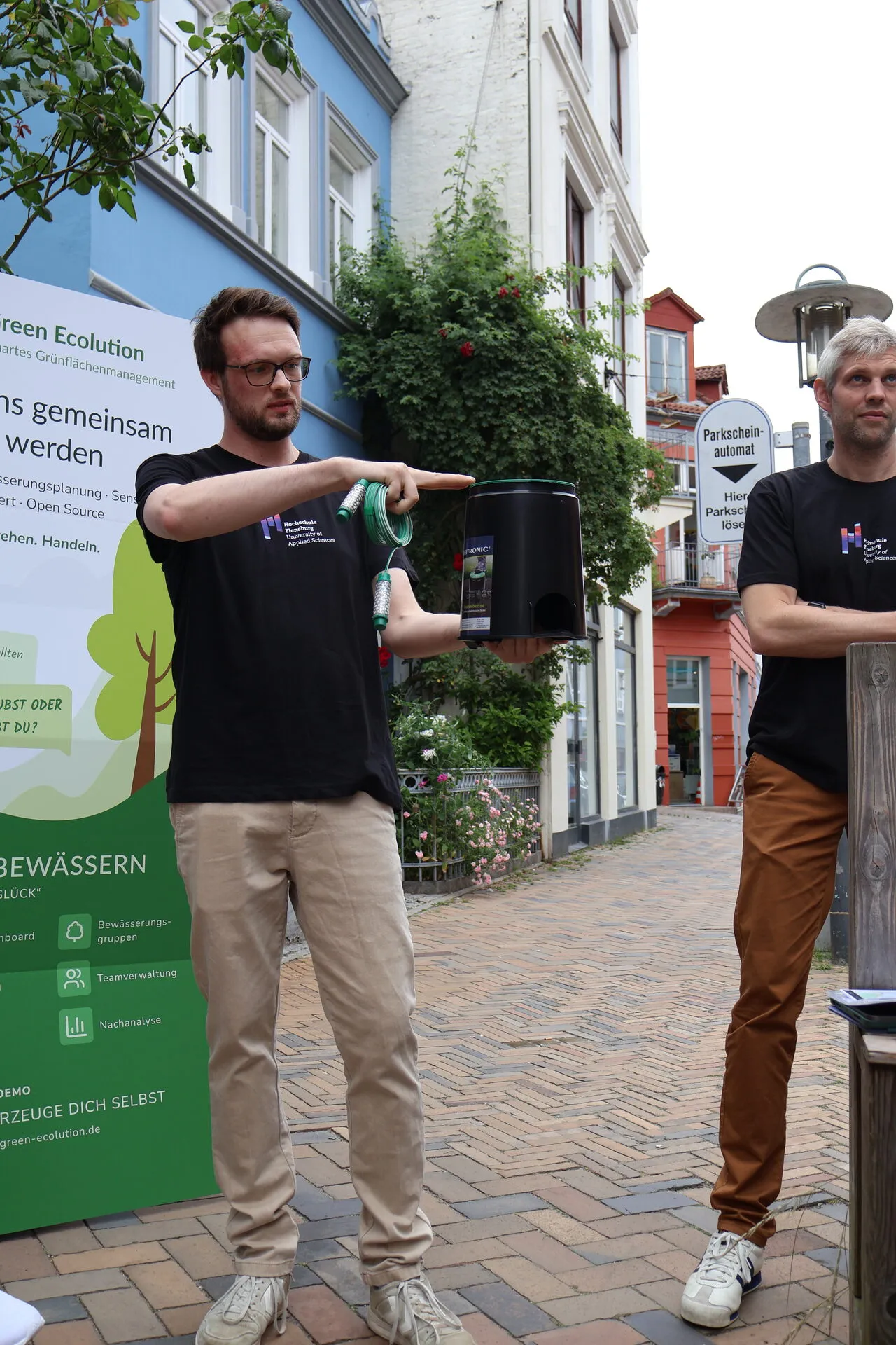 Cedrik Hoffmann hält vor dem Green-Ecolution-Rollup eine Bodendose und einen Watermark-Sensor hoch und erklärt beides, rechts neben ihm steht Simon Olberding