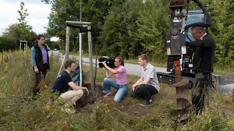 Ein Kamerateam filmt am Straßenrand den Einbau eines Bodenfeuchtesensors, daneben knien zwei Personen am Baum, rechts steht der Erdbohrer eines Baggers im Loch