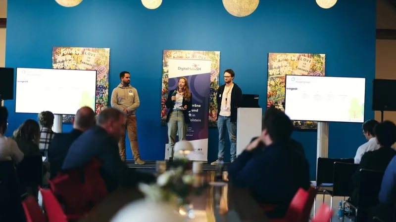 Drei Personen stellen Green Ecolution auf einer Bühne vor, zwischen zwei Bildschirmen steht ein Aufsteller des DigitalHub.SH